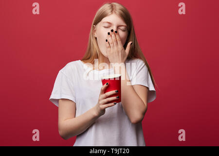 Faible jeune femme aux yeux clos du bâillement, fermant la bouche avec palm, essayant de ne pas s'endormir dans la matinée. Lundi,la photo en gros fond rouge isolé. Banque D'Images