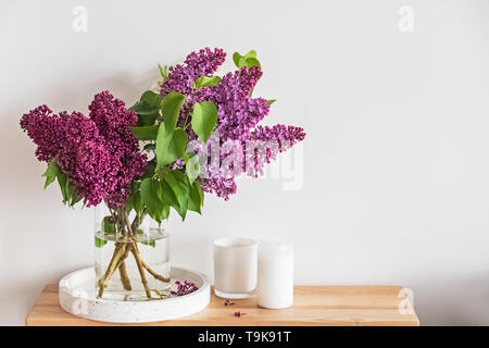 Bouquet de belles fleurs lilas debout dans un vase en verre sur le petit support en bois dans la salle de séjour avec des murs blancs Banque D'Images