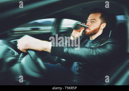 Un homme ivre au volant d'une voiture avec une bouteille d'alcool dans sa main. Homme d'une fiole de whisky de fer derrière le volant d'une voiture. Banque D'Images