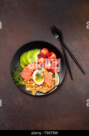 Gaufres de pommes de terre avec le saumon, d'oeufs, d'avocat et de tomates sur une plaque noire sur un fond sombre. Vue de dessus. Banque D'Images