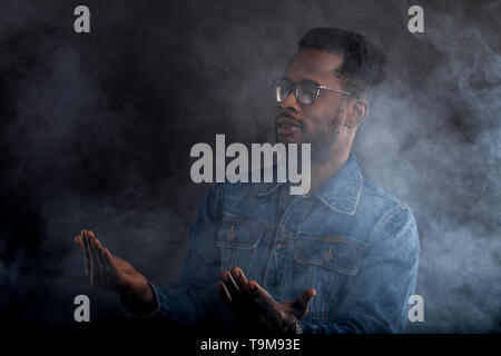 De quoi s'agit-il. Close up portrait of surpris handsome african american man à lunettes, habillé en passant l'article nuage de fumée blanche avec les paumes vers le haut, Banque D'Images