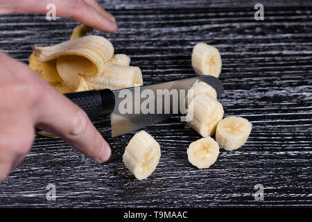 Un homme coupe un couteau de bananes en tranches sur une vieille table en bois noir. Le concept de la cuisson des plats sains rapide pour la perte de poids et la récupération du corps Banque D'Images