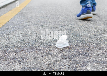 Tasse en plastique blanc froissé sur une route asphaltée, avec une bande jaune. Dans l'arrière-plan sont les jambes d'une personne qui a jeté la corbeille de départ Banque D'Images