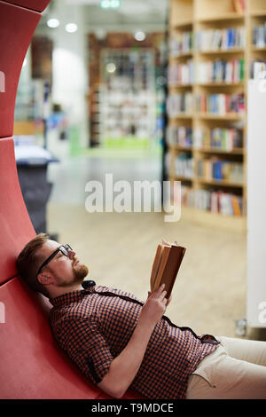 Bearded man reading book in library Banque D'Images