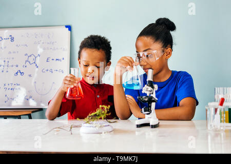 Cheerful black jolie enfants vêtements casual smart en séance à l'école laboratoire de chimie et holding flacons avec des liquides colorés Banque D'Images