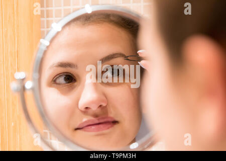 Belle femme avec des brucelles est plumer des sourcils tout en regardant dans le miroir dans la salle de bains. Beauty soin et bien-être matin concept. Banque D'Images