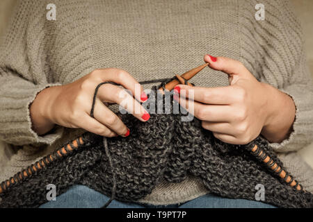 Girl with red nails le tricot de laine gris foncé avec des aiguilles en bois, assis sur une chaise, de l'artisanat avec de la laine Banque D'Images