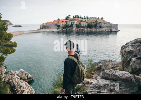 Touriste avec un sac à dos près de la mer. Il montre sa main sur l'île de Sveti Stefan au Monténégro. Banque D'Images