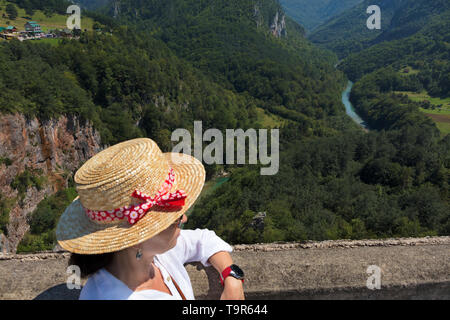 Une jeune femme en blouse blanche et chapeau de paille regarde la rivière de montagne qui s'écoule entre la gorge de pierre. Banque D'Images