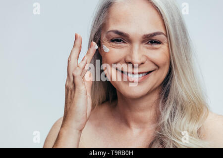 Smiling young caucasian woman crème anti de vieillissement sur le visage. Femme Senior woman applying moisturizer sur son visage contre l'arrière-plan gris. Banque D'Images