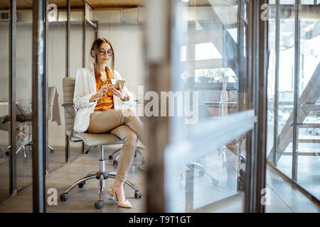 D'affaires élégante femme assise avec téléphone dans l'attente de la conférence dans la petite salle de réunion moderne Banque D'Images