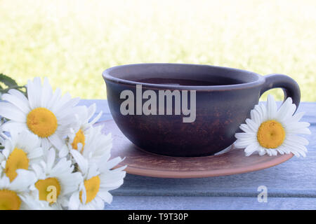 Une tasse de thé avec marguerites se dresse sur un tableau bleu sur fond d'herbe verte. Close-up. Banque D'Images