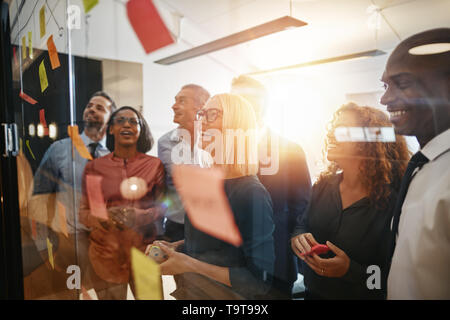 Groupe diversifié de smiling businesspeople avec brainstorming notes adhésives sur un mur de verre tout en travaillant dans un bureau moderne Banque D'Images