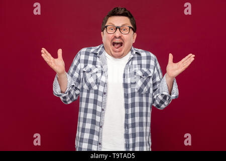 Portrait de surpris handsome middle aged man in casual chemise à carreaux et des lunettes debout avec les bras levés et à la caméra à l'INDO. Banque D'Images