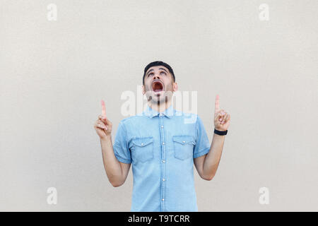 Portrait de choqué beau jeune homme barbu en chemise bleue en montrant le permanent jusqu'copyspace et à la face intérieure avec surprise. studio shot Banque D'Images