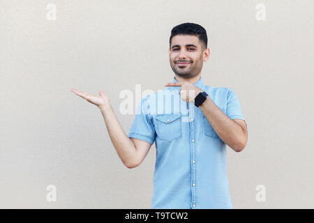 Portrait of happy satisfait beau jeune homme barbu en chemise bleue, tenant debout, pointant quelque chose et à la recherche à l'appareil photo avec sourire. Piscine st Banque D'Images