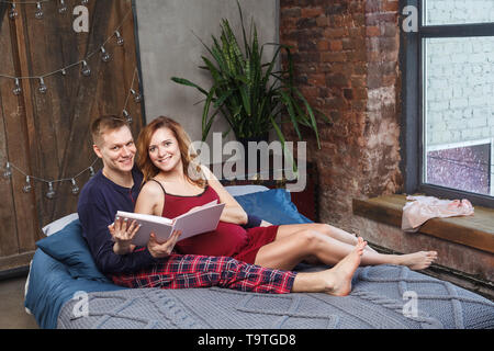 Portrait of happy young family se reposer dans la chambre à coucher, dans l'album parlons maintenant des vêtements et souriant tout en passant du temps ensemble au lit avec Banque D'Images