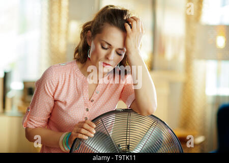Triste femme moderne dans la maison moderne en chaude journée d'été ensoleillée près de l'étage électrique ventilateur de pied souffrant de la chaleur de l'été. Banque D'Images
