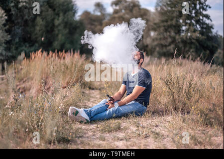 Fumeur barbu souffle beaucoup de fumée à l'aide de vape cigarette électronique dans la nature. Cigarette électronique. Banque D'Images