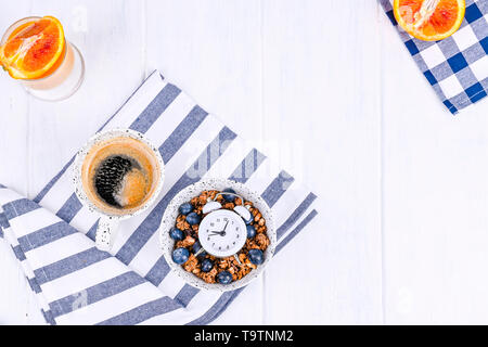 Muesli avec des bleuets dans une tasse, jus d'orange et aromatiques du café le matin. Le petit-déjeuner sur un fond en bois blanc et d'un réveil. Mise à plat. Copie Banque D'Images