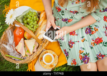 Femme sur le pique-nique se trouve sur la couverture jaune et maintenir le téléphone. Près de panier avec de la nourriture, des fruits et une tasse de thé. Banque D'Images