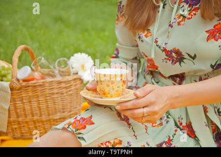 Femme sur le pique-nique se trouve sur la couverture jaune et est titulaire d'une tasse de thé ou café. Près de panier pique-nique avec de la nourriture, de fleurs et de fruits. Banque D'Images