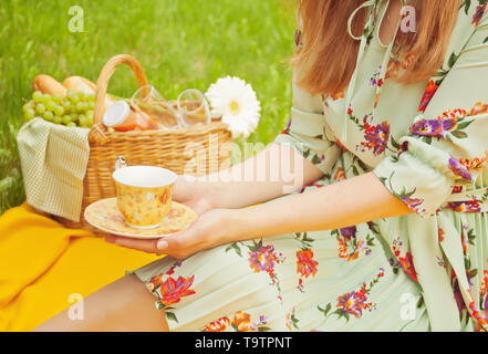 Femme sur le pique-nique se trouve sur la couverture jaune et est titulaire d'une tasse de thé ou café. Près de panier pique-nique avec de la nourriture, de fleurs et de fruits. Banque D'Images