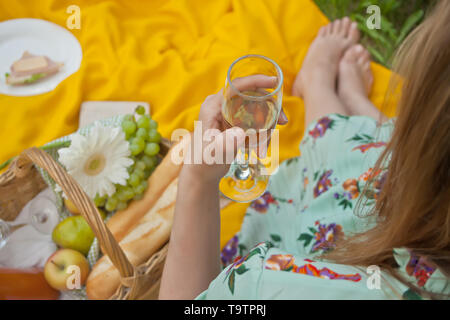 Femme sur le pique-nique se trouve sur la couverture jaune et est titulaire d'un verre de vin. Près de panier pique-nique avec de la nourriture, de fleurs et de fruits. Banque D'Images