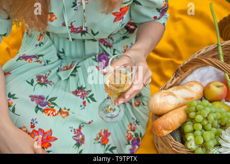 Femme sur le pique-nique se trouve sur la couverture jaune et est titulaire d'un verre de vin. Près de panier pique-nique avec de la nourriture, de fleurs et de fruits. Banque D'Images