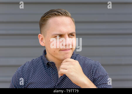 Jeune homme attentionné avec sa main à son menton fixant sur le côté à la recherche de ses lèvres en concentration devant un mur gris with copy space Banque D'Images