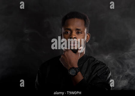 Portrait de teint foncé homme sérieux holding chin à la caméra à mystérieusement, vêtus de noir, veste coupe-vent wristwatces, debout contre Banque D'Images
