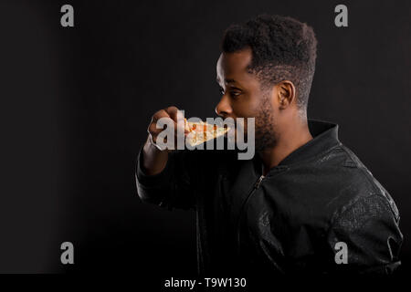 Closeup portrait of african american non rasé faim guy avec morceau de pizza savoureuse dans sa main, eating fast food isolé sur fond noir. Unhealt Banque D'Images