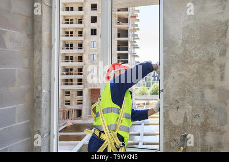 Un travailleur dans un casque orange installe un double-vitrage dans une nouvelle maison, dans un contexte de construction. Banque D'Images