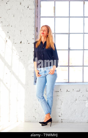 Toute la longueur de balle belle blonde woman wearing shirt et jeans tout en se tenant à la fenêtre et la rêverie. Banque D'Images