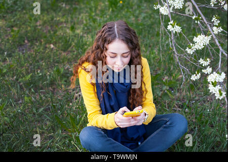Jeune femme souriant assis dans un jardin fleuri et écrit sur téléphone mobile. Cerisier en fleurs. Portrait de jeune fille Banque D'Images