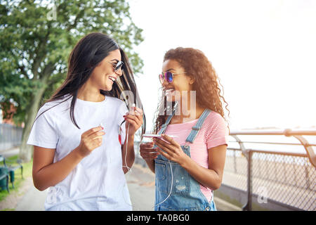 Deux jeunes copines sourire marcher ensemble vers le bas une promenade dans l'été à l'écoute de la musique sur une paire d'écouteurs Banque D'Images