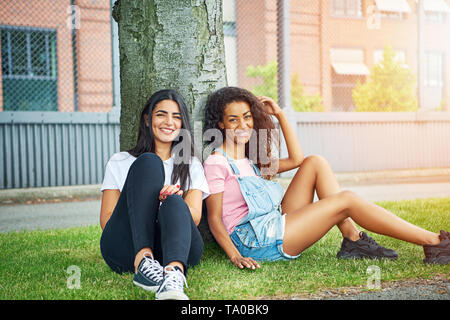 Deux jeunes femmes attrayantes tout en souriant, assis sur une pelouse, se penchant en arrière sur un tronc d'arbre sur une journée ensoleillée Banque D'Images