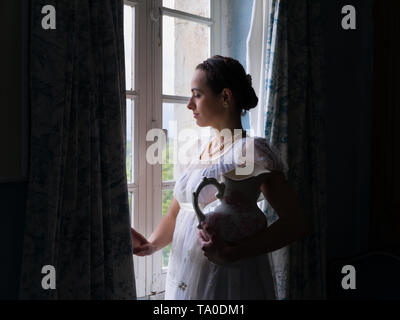 Jeune femme en robe regency authentique près d'une fenêtre d'un intérieur classique Banque D'Images