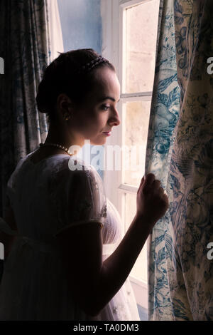 Jeune femme en robe regency authentique près d'une fenêtre d'un intérieur classique Banque D'Images