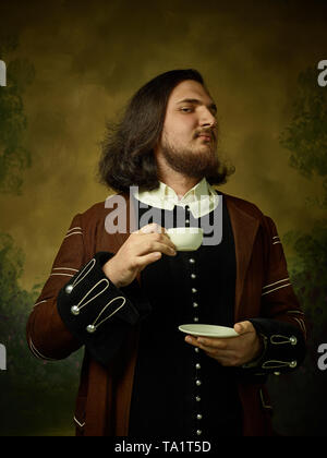 Jeune homme comme un chevalier médiéval sombre sur fond de studio. Portrait en bas de la clé modèle masculin en costume rétro. Boire le café du matin. Les émotions humaines, Banque D'Images