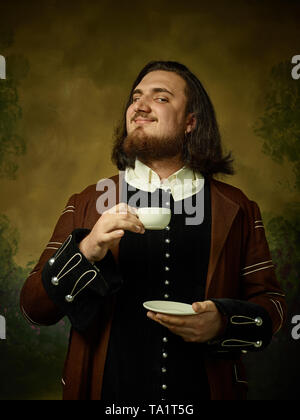 Jeune homme comme un chevalier médiéval sombre sur fond de studio. Portrait en bas de la clé modèle masculin en costume rétro. Boire le café du matin. Les émotions humaines, Banque D'Images