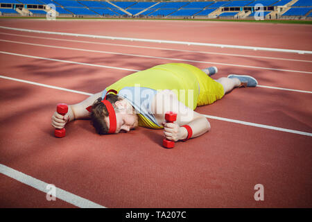 Le gros homme dans les vêtements de sport est fatigué et dort avec haltères en ses mains sur la piste du sport dans le stade. Banque D'Images