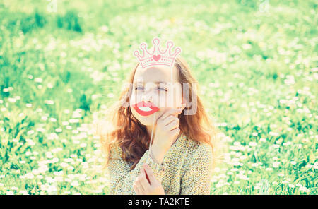 L'humour reine concept. Fille est assise sur l'herbe à grassplot, fond vert. Fille sur le visage rieur qui passent à l'extérieur loisirs. Enfant posant avec un sourire en carton les lèvres et couronne pour séance photos à Meadow. Banque D'Images