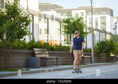 Élégante sportive plussize girl à lunettes monte un longboard le long du chemin d'un tracksporty cours des arbres et de bancs Banque D'Images