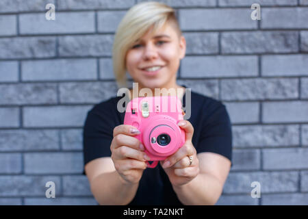 Grande élégante fille dans un t-shirt noir sourit et prend des photos sur un appareil photo rose contre le mur Banque D'Images