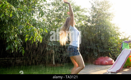 Portrait of beautiful smiling woman in vêtements humides bénéficiant d'pluie chaude au jardin à l'arrière de la chambre à coucher du soleil. Girl le jeu et le plaisir en plein air au Banque D'Images