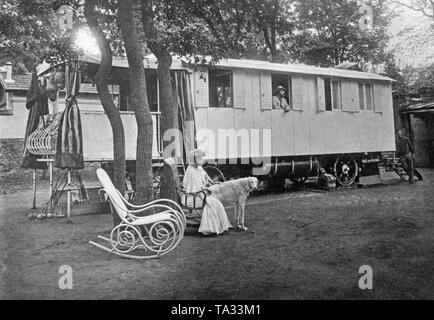 Camping de luxe, historique ( 'MOTOR' des voitures de tourisme), en face d'elle une femme est assise dans un fauteuil à bascule, à côté d'elle est un chien. Un homme est à la fenêtre de la caravane et est à l'extérieur. Banque D'Images