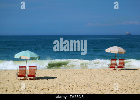 Ensemble de deux chaises de plage rouge et bleu un parasol de plage sur la plage de sable en face du fracas des vagues et l'océan bleu vif Banque D'Images