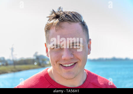 Avant Close-up portrait of smiling caucasian type en t-shirt et coupe courte, debout à l'extérieur avec de l'eau d'un lac ou de la rivière en arrière-plan Banque D'Images