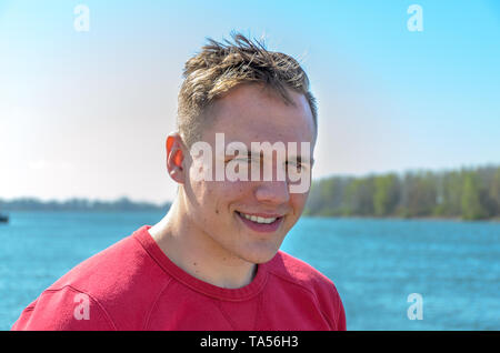 Demi-tour buste portrait d'un jeune gars en t-shirt rouge, debout à l'extérieur, sur le lac ou la rivière. Looking at camera and smiling Banque D'Images